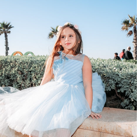  little girl sitting and smiling outdoors in a blue dress and flower crown like the ones they sell at Asdelia Mae Children's Clothing Boutique